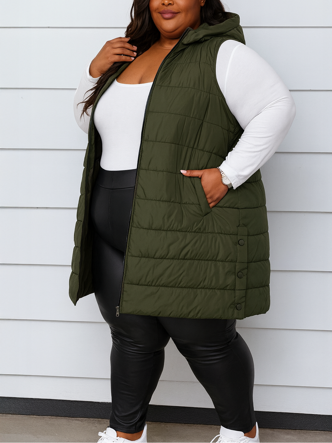 Person wearing an olive green puffer vest over a white long-sleeve shirt and black leggings against a light gray background.