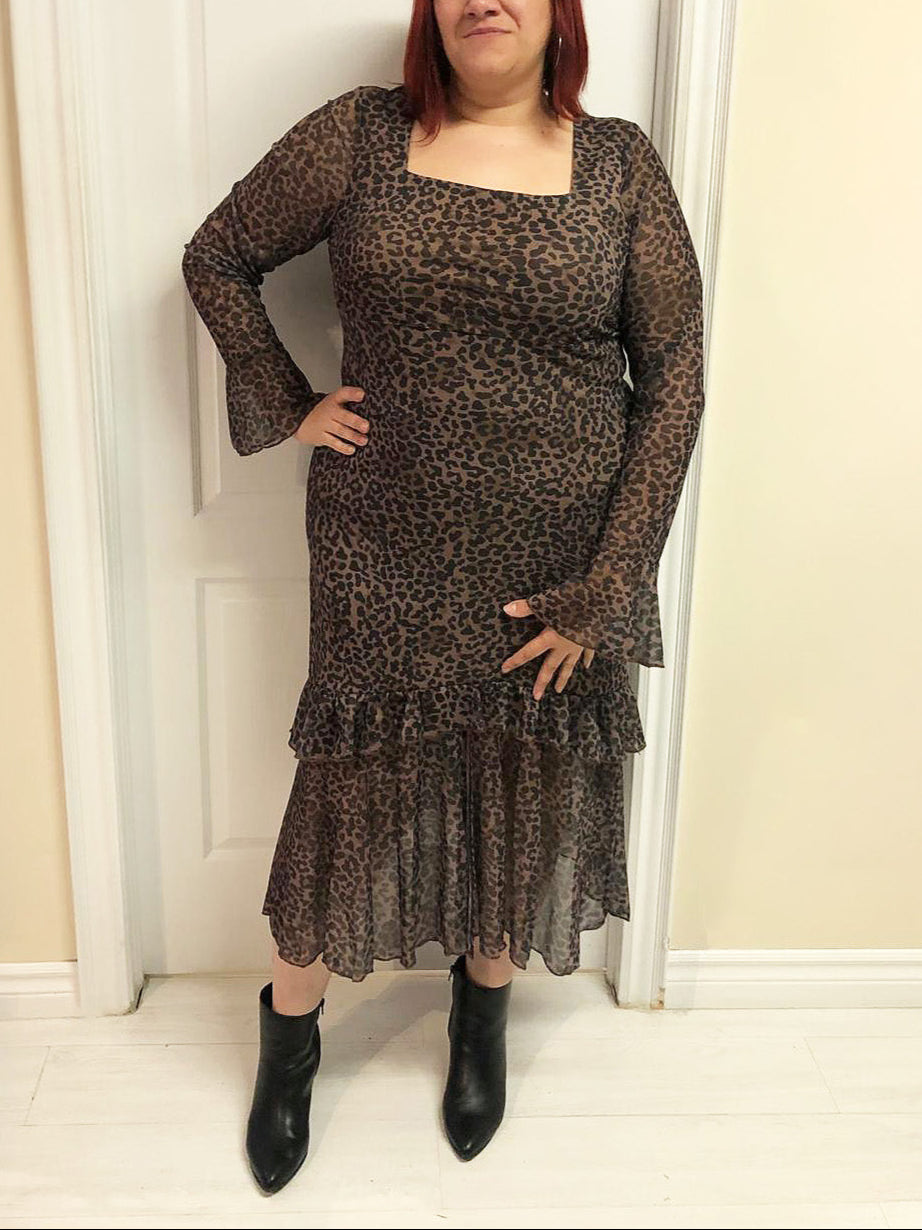 Plus size woman wearing a leopard print dress with long sleeves and ruffled hem, standing against a neutral wall.
