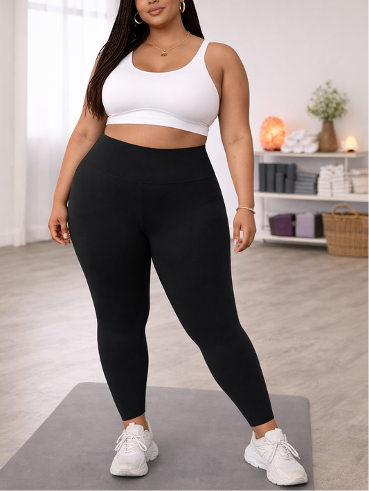 Woman in a white sports bra and black leggings standing on a yoga mat in a room with a shelf in the background.