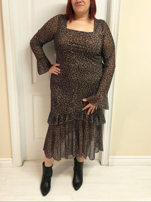 Plus size woman wearing a leopard print dress with long sleeves and ruffled hem, standing against a neutral wall.