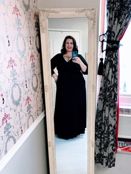 Person wearing a black dress standing in front of a full-length mirror in a room with decorative wallpaper and curtains.